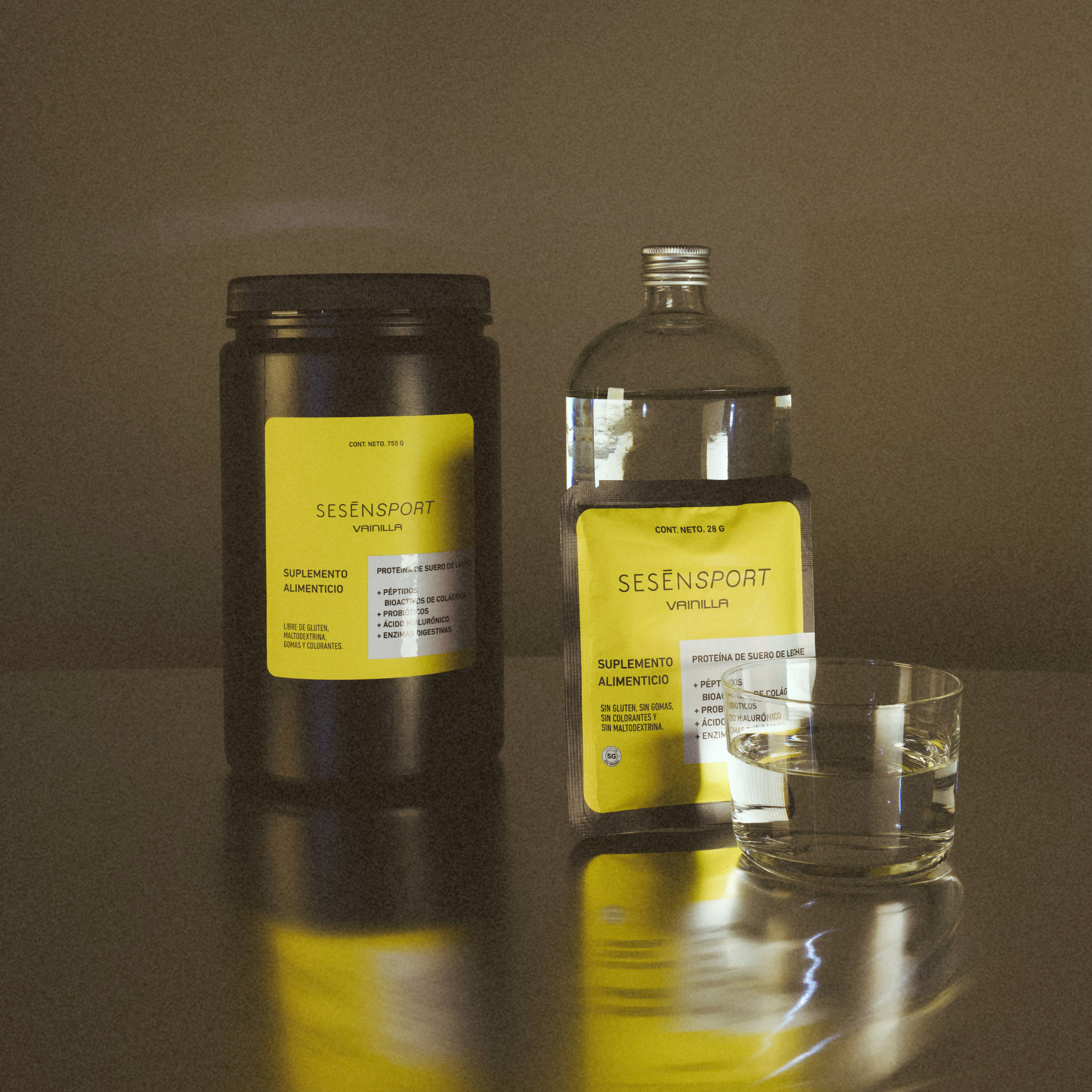 Proteína whey sabor vainilla en envases con etiqueta amarilla, acompañada de un vaso con líquido sobre una superficie reflectante.