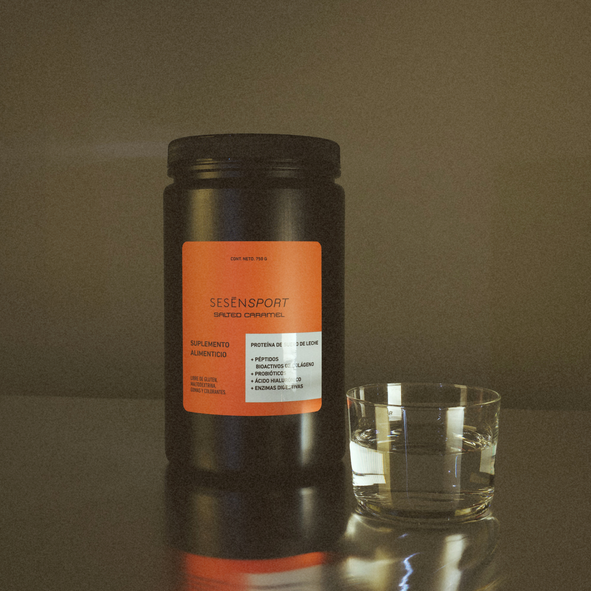 Proteína whey sabor salted caramel en envase negro con etiqueta naranja, acompañada de un vaso con líquido sobre una superficie reflectante.